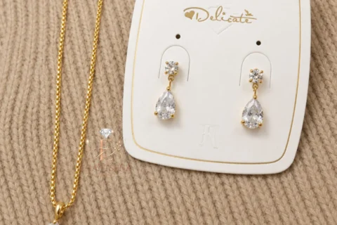 Ensemble de bijoux doré pour femme comprenant un collier avec pendentif en forme de goutte et des boucles d'oreilles pendantes assorties ornées de pierres transparentes.