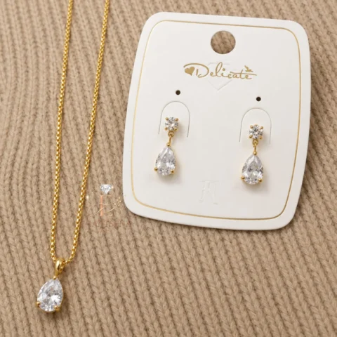 Ensemble de bijoux doré pour femme comprenant un collier avec pendentif en forme de goutte et des boucles d'oreilles pendantes assorties ornées de pierres transparentes.